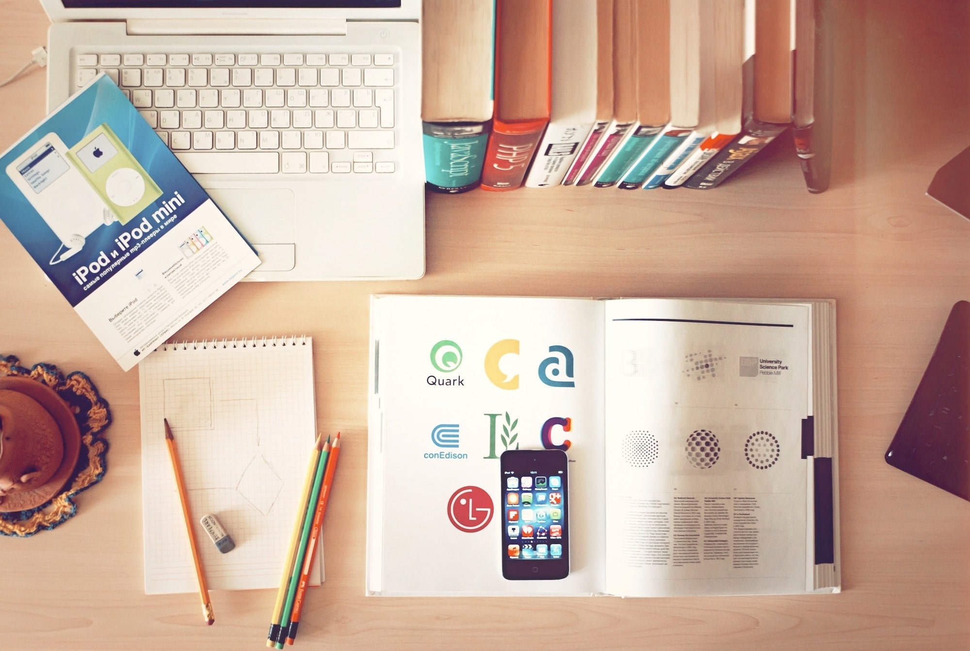 Overhead view of a desk with a computer, several books, an open magazine with a cell phone laying on it, several pencils, and an iPod mini user manual